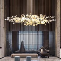 A luxurious lobby featuring a striking horizontal chandelier with illuminated leaf-shaped elements. The fixture hangs against a backdrop of vertical dark stripes. Below, modern seating includes a long bench and sculptural chairs. Marble columns frame the space, while a textured metal screen adds depth. The room blends organic shapes with contemporary design, creating an atmosphere of sophisticated elegance.