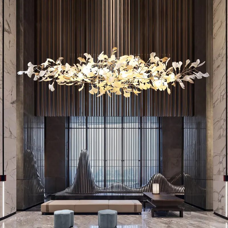 A luxurious lobby featuring a striking horizontal chandelier with illuminated leaf-shaped elements. The fixture hangs against a backdrop of vertical dark stripes. Below, modern seating includes a long bench and sculptural chairs. Marble columns frame the space, while a textured metal screen adds depth. The room blends organic shapes with contemporary design, creating an atmosphere of sophisticated elegance.