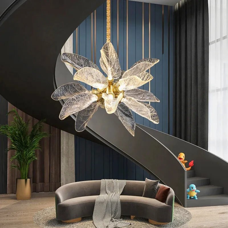 A contemporary living area featuring a striking glass leaf chandelier. The chandelier, with its illuminated translucent leaves, hangs above a curved gray sofa. A dramatic curved staircase frames the scene, with colorful toy figures on its steps. The background showcases a blue striped wall. A potted plant adds a touch of nature. The space blends modern design elements with playful accents, creating an inviting and stylish atmosphere.