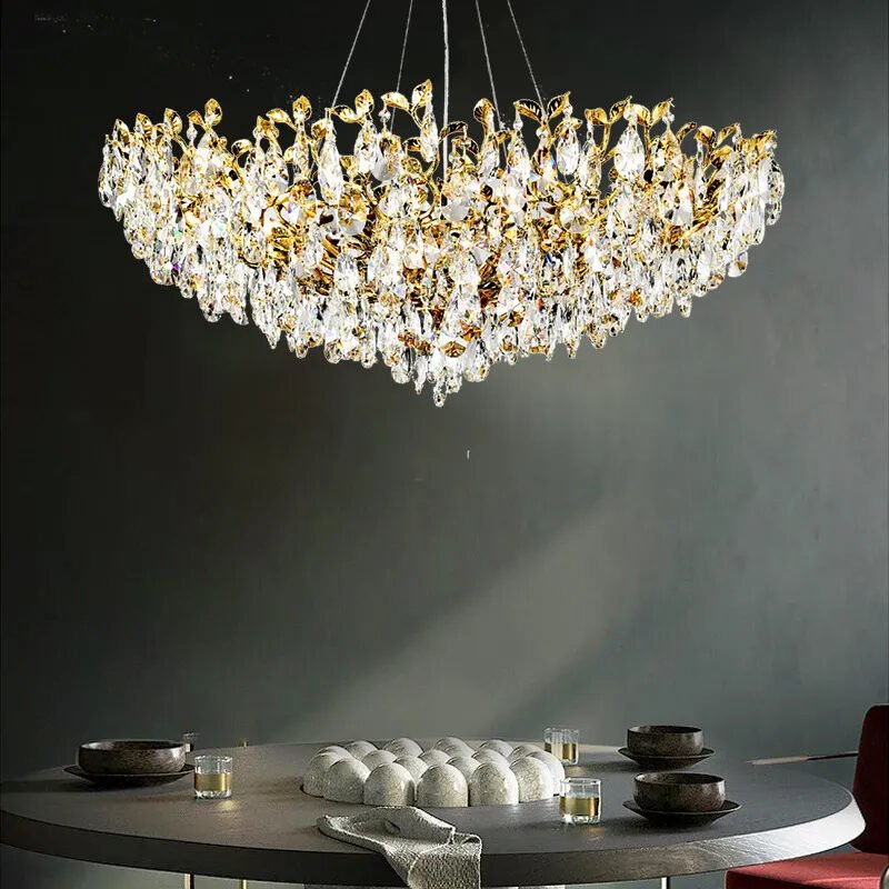 A grand gold and crystal chandelier illuminates a modern dining setup. The chandelier features intricate gold branches adorned with numerous teardrop-shaped crystal pendants. Below, a round table is set with dark plates, glasses, and a striking white circular cake. Dramatic lighting casts shadows on the dark wall, creating an atmosphere of sophistication and luxury.