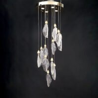 An elegant branch chandelier featuring translucent glass leaves suspended at varying lengths from a circular gold-toned base. Each leaf-shaped glass element is delicately textured to mimic real foliage and is topped with a small, ornate gold cap. The chandelier creates a cascading effect against a dark background, highlighting the intricate details of each glass leaf. LED spotlights integrated into the circular base provide additional illumination, making the glass leaves appear to glow. This modern branch chandelier beautifully combines nature-inspired forms with luxurious materials, creating a stunning interplay of light and shadow that would serve as a captivating focal point in any space.