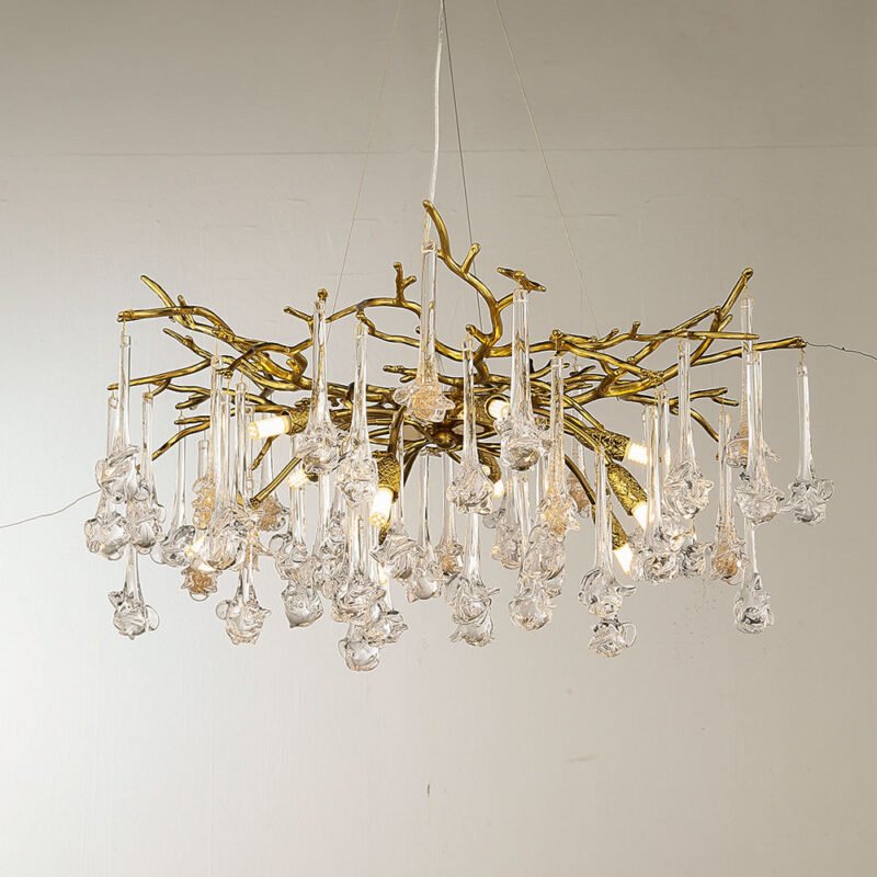 A luxurious chandelier suspended against a light background, showcasing a harmonious blend of gold-toned metalwork and clear glass elements. The fixture's core structure consists of intricately designed gold-plated branches that twist and intertwine, creating an organic, tree-like silhouette. These golden branches extend outward in various directions, forming a wide, sprawling canopy. Hanging from the golden branches are numerous clear glass pendants, each with a unique, elongated shape. These glass elements feature long, slender stems that transition into more elaborate, bulbous forms at their ends, reminiscent of abstract flowers or icicles. The glass pendants vary in length, creating a dynamic, cascading effect that adds depth and dimension to the overall design. The chandelier is illuminated from within, with several LED lights visible among the branches. These lights cast a warm glow that highlights both the metallic gold tones of the branches and the translucent quality of the glass pendants. The light refracts and reflects through the glass elements, creating a dazzling display of sparkles and subtle rainbow effects. The piece is suspended by thin wires from the ceiling, giving it a floating appearance. The contrast between the warm gold of the branches and the cool, clear glass of the pendants creates a striking visual balance. This chandelier beautifully combines natural, organic forms with luxurious materials and modern lighting technology, resulting in a piece that is both a functional light source and a stunning sculptural element. It would serve as an impressive focal point in contemporary or transitional interior spaces, particularly suited for dining areas, grand entryways, or upscale living rooms.