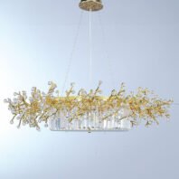 An elegant oval-shaped chandelier suspended against a light background. The fixture features a central oval base composed of vertically arranged crystal prisms, creating a luminous, curtain-like effect. Above this base, an intricate network of gold-plated branches extends outward, reminiscent of a golden coral reef or a stylized tree canopy. These branches are adorned with numerous faceted crystal orbs of varying sizes, resembling sparkling dewdrops or blossoms. The crystals catch and refract light brilliantly, adding a dazzling dimension to the piece. The gold branches create an organic, flowing silhouette that contrasts beautifully with the geometric precision of the crystal prism base. The chandelier is suspended by thin wires from a circular gold-toned ceiling mount with a textured, ornate design. The light background allows the intricate details of the chandelier to stand out clearly, emphasizing the interplay between the warm gold tones and the clear, sparkling crystals. This chandelier masterfully combines nature-inspired design with luxurious materials, resulting in a piece that is both a functional light source and an exquisite work of art. It would serve as a stunning focal point in any high-end interior space, particularly suited for dining areas or grand entryways.