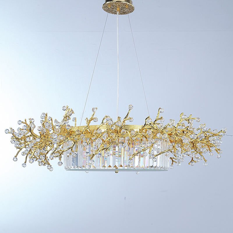 An elegant oval-shaped chandelier suspended against a light background. The fixture features a central oval base composed of vertically arranged crystal prisms, creating a luminous, curtain-like effect. Above this base, an intricate network of gold-plated branches extends outward, reminiscent of a golden coral reef or a stylized tree canopy. These branches are adorned with numerous faceted crystal orbs of varying sizes, resembling sparkling dewdrops or blossoms. The crystals catch and refract light brilliantly, adding a dazzling dimension to the piece. The gold branches create an organic, flowing silhouette that contrasts beautifully with the geometric precision of the crystal prism base. The chandelier is suspended by thin wires from a circular gold-toned ceiling mount with a textured, ornate design. The light background allows the intricate details of the chandelier to stand out clearly, emphasizing the interplay between the warm gold tones and the clear, sparkling crystals. This chandelier masterfully combines nature-inspired design with luxurious materials, resulting in a piece that is both a functional light source and an exquisite work of art. It would serve as a stunning focal point in any high-end interior space, particularly suited for dining areas or grand entryways.