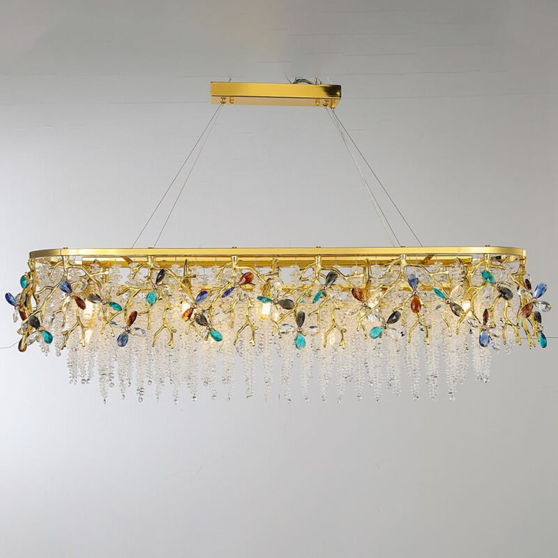 An opulent oval-shaped chandelier suspended against a light background, showcasing a stunning combination of gold, clear crystals, and multi-colored gemstone-like accents. The fixture features a polished gold oval frame, hanging from a rectangular gold ceiling mount via thin cables. Intricate golden branches extend from this frame, adorned with vibrant crystal flowers in various colors including blue, turquoise, amber, and clear. These colorful crystal accents provide eye-catching pops of color against the predominantly gold and clear crystal design. The most striking feature is the abundant cascade of clear crystal strands hanging from the gold frame, creating a shimmering curtain effect. These crystal strands are arranged in varying lengths, giving the chandelier a waterfall-like appearance. The clear crystals catch and refract light, producing a dazzling sparkle. Several small light bulbs are visible within the structure, promising a warm, inviting glow when lit. This chandelier masterfully combines the warmth of gold with the cool brilliance of crystals and the vibrancy of colored gemstones, resulting in a piece that is both a functional light source and an exquisite work of art. It's particularly well-suited for long dining tables, expansive living areas, or as a centerpiece in grand entryways, capable of making a dramatic statement in any luxurious interior space.