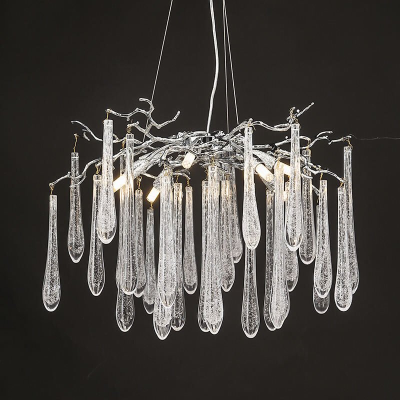 A mesmerizing branch chandelier featuring a network of silver-toned metallic branches that resemble bare winter trees. The chandelier's structure is crafted from polished chrome or nickel, creating an intricate, organic silhouette against a dark background. Suspended from the silver branches are numerous elongated crystal pendants of varying lengths, reminiscent of icicles or frozen raindrops. These transparent crystal elements have a textured, bubbled surface that enhances light refraction. Warm LED lights are nestled within the branches, casting a soft glow that illuminates the crystal droplets from within, creating a magical, wintry atmosphere. This branch chandelier combines the stark beauty of winter trees with the sparkle of ice crystals, perfect for adding a touch of enchantment to modern or contemporary interiors.