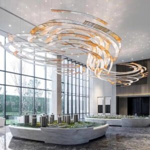 A stunning large chandelier dominates a contemporary hotel lobby, showcasing innovative custom lighting design. This big chandelier features graceful, ribbon-like metallic bands that swirl and intertwine, creating a dynamic sculptural effect. The chandelier's warm golden and silver hues contrast beautifully with the lobby's bright, airy atmosphere. Floor-to-ceiling windows flood the space with natural light, while a central display showcases miniature building models. Suspended small lights above create a starry sky effect. This unique hotel chandelier transforms the expansive lobby into a captivating blend of artistry and functionality, exemplifying how bespoke lighting can elevate luxury hospitality spaces to new heights of sophistication and visual appeal.