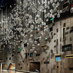 A stunning large chandelier dominates a unique hotel interior, showcasing innovative custom lighting design. This big chandelier features hundreds of suspended crystal cubes, creating an illusion of a crystalline rain or icicles frozen in mid-air. The chandelier elements are hung at varying heights, casting a soft, ethereal glow across the space. Below, the walls exhibit an avant-garde design with an array of irregularly placed windows and recesses in various shapes, sizes, and colors, including touches of green and gold. The concrete walls provide a stark, industrial contrast to the delicate chandelier. At the bottom of the image, a glimpse of the floor reveals a colorful mosaic or artwork, adding another layer of visual interest to this captivating hotel interior that seamlessly blends luxury with contemporary artistic vision.