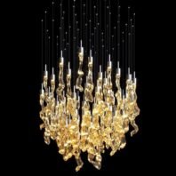 A magnificent large chandelier showcases innovative custom lighting design for luxury hotels. This big chandelier features a dense cluster of golden, organically-shaped glass elements suspended at varying lengths, creating a mesmerizing waterfall effect. Each piece glows warmly, resembling molten gold or amber crystals. Thin silver wires extend upward from the glass elements, topped with tiny LED lights, adding a starry canopy effect against the black background. This unique hotel chandelier transforms the space with its dynamic interplay of light and form, offering a stunning focal point that embodies both opulence and contemporary artistry.