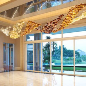 A magnificent big chandelier adorns a luxurious hotel lobby, showcasing exquisite custom lighting design. The large chandelier features a flowing wave pattern of golden glass elements, transitioning from deep amber to light gold. Above, a recessed ceiling with blue accent lighting adds depth. Floor-to-ceiling windows reveal a stunning tropical landscape with palm trees and a serene pool, creating a seamless blend of interior opulence and exterior paradise.