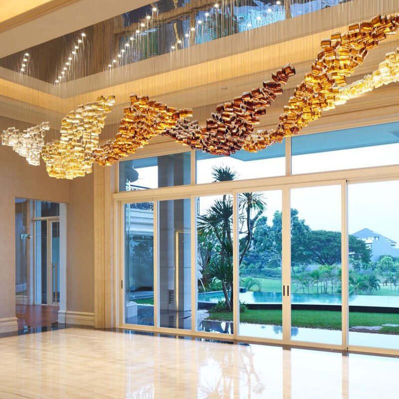 A magnificent big chandelier adorns a luxurious hotel lobby, showcasing exquisite custom lighting design. The large chandelier features a flowing wave pattern of golden glass elements, transitioning from deep amber to light gold. Above, a recessed ceiling with blue accent lighting adds depth. Floor-to-ceiling windows reveal a stunning tropical landscape with palm trees and a serene pool, creating a seamless blend of interior opulence and exterior paradise.