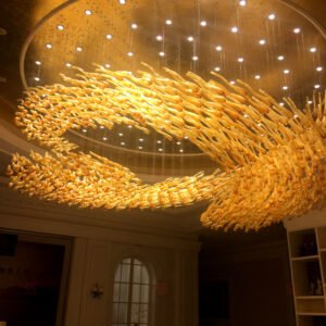 A large custom lighting hotel chandelier dominates a circular ceiling recess. This big chandelier features thousands of golden, elongated glass elements arranged in a dynamic wave-like pattern. The warm, glowing pieces resemble a swirling school of fish or a golden tsunami, creating a sense of fluid motion. Interspersed white spotlights add depth and sparkle to the installation. This unique custom lighting design transforms the hotel space into a mesmerizing, luxurious environment, capturing the essence of flowing water or wind-swept sand dunes in a stunning, abstract form.