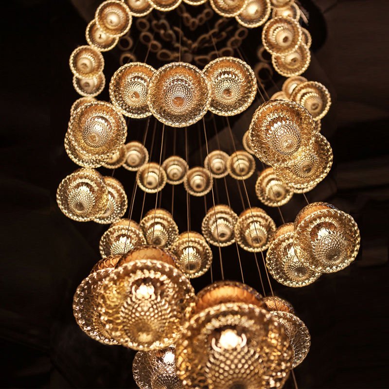 A stunning custom lighting installation featuring a large hotel chandelier composed of multiple golden, disc-shaped elements. This big chandelier showcases an array of intricately patterned glass or crystal discs of varying sizes, each emitting a warm, golden glow. The custom lighting piece is arranged in a cascading formation, with smaller discs forming a circular pattern at the top and larger, more elaborately designed discs suspended lower. Thin wires support each element, creating an ethereal, floating effect. The chandelier's luxurious design and radiant illumination make it a perfect centerpiece for upscale spaces, blending artistry with functionality.