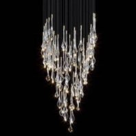 A stunning custom lighting installation featuring a large chandelier with a dramatic V-shaped design. This hotel chandelier showcases numerous crystal-like glass elements suspended at varying lengths on thin wires against a dark background. The glass pieces range from elongated teardrops to twisted, abstract forms, each capturing and refracting light brilliantly. The chandelier creates a cascading effect, with the glass elements densely clustered at the top and gracefully tapering towards a point at the bottom. Each glass piece is illuminated, emitting a warm, sparkling glow that ranges from bright white to soft amber hues. The arrangement of the glass elements creates a sense of movement, as if light is flowing downward. This big chandelier offers a perfect blend of elegance and contemporary design, ideal for creating a captivating focal point in luxurious spaces.