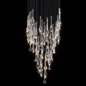 A stunning custom lighting installation featuring a large chandelier with a dramatic V-shaped design. This hotel chandelier showcases numerous crystal-like glass elements suspended at varying lengths on thin wires against a dark background. The glass pieces range from elongated teardrops to twisted, abstract forms, each capturing and refracting light brilliantly. The chandelier creates a cascading effect, with the glass elements densely clustered at the top and gracefully tapering towards a point at the bottom. Each glass piece is illuminated, emitting a warm, sparkling glow that ranges from bright white to soft amber hues. The arrangement of the glass elements creates a sense of movement, as if light is flowing downward. This big chandelier offers a perfect blend of elegance and contemporary design, ideal for creating a captivating focal point in luxurious spaces.