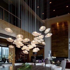 A stunning custom lighting installation dominates the high-ceilinged space of a modern, luxurious loft. Multiple organically shaped glass elements, resembling ethereal clouds or abstract pebbles, are suspended at varying heights from thin wires. Each translucent glass piece emits a soft, warm glow, creating a floating constellation of light. The installation is clustered primarily above a large dining table, but extends throughout the open-plan living area. The loft features a mix of contemporary and natural elements. A dark wooden ceiling contrasts with lighter wood paneling on the walls. Floor-to-ceiling windows offer a view of a cityscape, while allowing natural light to complement the ambient lighting. The space is furnished with modern, neutral-toned seating and accented with purple throw pillows. A tropical leaf on the dining table adds a touch of greenery. This innovative lighting design serves as a captivating focal point, transforming the loft into a sophisticated, atmospheric space that seamlessly blends artistic lighting with high-end interior design. The installation creates a sense of wonder and lightness, perfectly balancing the loft's modern architectural elements with organic, cloud-like forms.