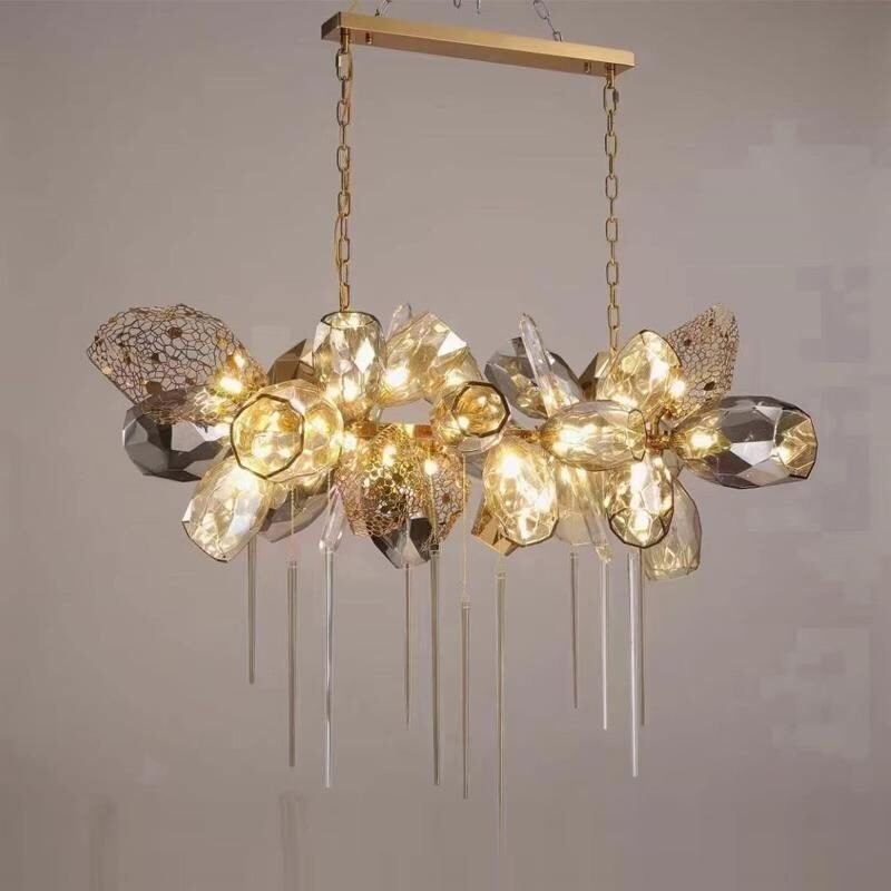 A stunning linear chandelier showcasing innovative custom lighting design. The fixture features a horizontal gold-toned metal bar suspended by two chains. From this bar hangs a cluster of diverse geometric crystal-like elements in various shapes, sizes, and textures. The palette includes clear, smoky gray, and champagne-colored pieces, with some having intricate filigree or honeycomb patterns. The arrangement is asymmetrical and organic, creating a cloud-like formation. What sets this image apart is the warm, golden illumination emanating from within the crystal elements, causing them to glow brilliantly and creating a magical, ethereal effect. Beneath the main cluster, multiple slender crystal rods hang vertically at varying lengths, adding a delicate, icicle-like quality to the design. This luxurious custom lighting piece would serve as a captivating focal point over a dining table or in a grand entryway, perfect for those seeking a blend of contemporary design and radiant, warm illumination in their lighting.