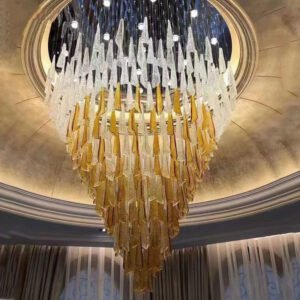 A spectacular custom chandelier hangs from a domed ceiling, showcasing an innovative lighting design. The chandelier features numerous elongated crystal-like elements arranged in a cascading conical shape. The top layers consist of clear or white crystals, gradually transitioning to golden hues towards the bottom, creating a stunning ombre effect. The chandelier is set within a circular recess in the dome, which is adorned with concentric rings and has a dark blue background, giving the illusion of a night sky. Small globe lights are interspersed among the crystal elements, adding to the celestial feel. The surrounding dome is finished in a warm, metallic gold color with subtle texturing, enhancing the luxurious ambiance. This opulent lighting installation perfectly blends contemporary design with classic elegance, ideal for upscale hotels or grand spaces seeking a breathtaking focal point.