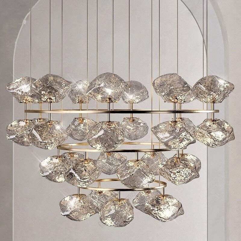 A stunning large hotel chandelier showcasing innovative custom lighting design. This big chandelier features four tiers of irregular, cloud-like glass elements suspended from gold-toned metal rings. Each crystalline glass piece is brilliantly illuminated, creating a dazzling display reminiscent of ice formations or rough-cut diamonds. The chandelier hangs within an arched alcove with light gray paneled walls, enhancing its ethereal presence. This luxurious custom lighting installation serves as a captivating focal point, perfect for upscale hotels seeking an elegant, modern centerpiece that combines contemporary design with timeless glamour and transforms the space with its celestial glow.