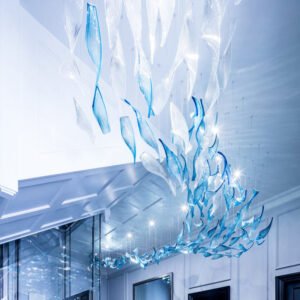 A breathtaking large hotel chandelier showcasing innovative custom lighting design. This big chandelier features numerous blue and clear glass elements shaped like abstract waves or leaves, suspended from the ceiling in a flowing, cascading pattern. The glass pieces appear to float weightlessly, creating a dynamic, undulating form that stretches along the hallway. Each element is delicately illuminated, casting a soft, ethereal glow that reflects off nearby surfaces. This stunning custom lighting installation transforms the modern, white-walled space into a mesmerizing underwater-like environment, perfect for luxury hotels seeking a unique, immersive centerpiece that captivates guests with its fluid beauty and serene ambiance.