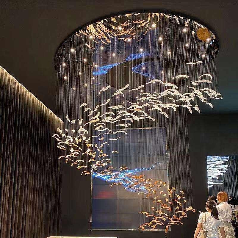 An awe-inspiring large hotel chandelier showcasing innovative custom lighting design. This big chandelier features hundreds of metallic feather-shaped elements suspended from a circular ceiling mount, creating a cascading effect. Interwoven with thin wires and warm white LED lights, the chandelier is accented by striking blue LED streams, mimicking a cosmic river. The gradient from silver to golden feathers adds depth and movement. Two observers stand beneath, emphasizing the chandelier's grand scale and mesmerizing presence in the hotel space.