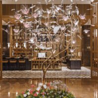 A breathtaking large custom chandelier in a luxury hotel lobby, designed as an enchanted tree. This big chandelier features a golden, intricately twisted trunk with branches extending outwards. Suspended from the branches are numerous pink and clear glass elements resembling flowers and leaves, creating a magical, floating effect. The chandelier spans from floor to ceiling, dominating the space with its ethereal beauty. Illuminated glass blossoms add a warm, inviting glow. Below, a floral arrangement complements the tree design. The chandelier is set against a backdrop of wooden panels and geometric flooring, seamlessly blending with the hotel's elegant interior, showcasing the pinnacle of custom lighting artistry.