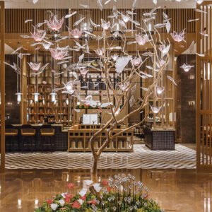 A breathtaking large custom chandelier in a luxury hotel lobby, designed as an enchanted tree. This big chandelier features a golden, intricately twisted trunk with branches extending outwards. Suspended from the branches are numerous pink and clear glass elements resembling flowers and leaves, creating a magical, floating effect. The chandelier spans from floor to ceiling, dominating the space with its ethereal beauty. Illuminated glass blossoms add a warm, inviting glow. Below, a floral arrangement complements the tree design. The chandelier is set against a backdrop of wooden panels and geometric flooring, seamlessly blending with the hotel's elegant interior, showcasing the pinnacle of custom lighting artistry.