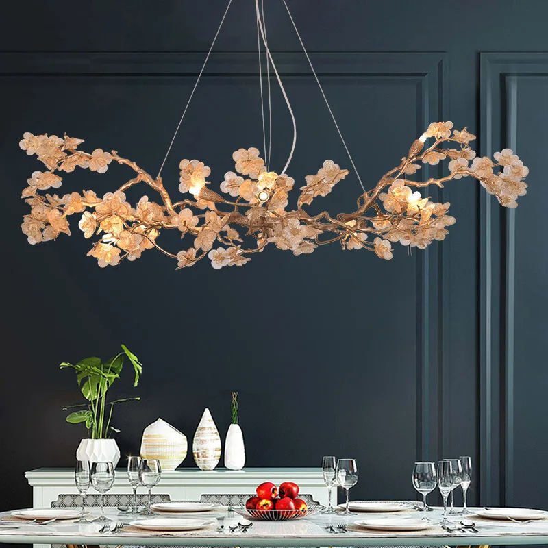 A luxurious dining room featuring a stunning gold-toned floral chandelier against a dark blue-gray wall. The chandelier has an organic, branch-like design adorned with numerous glass flowers, creating a delicate and ethereal appearance. It's suspended above a white dining table set with plates, wine glasses, and a bowl of red apples. The table also displays decorative vases and a small potted plant. The dark wall behind has classic molding details, adding a touch of traditional elegance to the modern chandelier design. The warm glow from the chandelier's lights contrasts beautifully with the cool-toned wall, creating an inviting and sophisticated dining atmosphere.