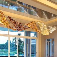 A stunning large chandelier dominates a hotel lobby, showcasing custom lighting design. The big chandelier features a wave-like pattern of golden glass elements, transitioning from deep amber to light gold. Above, a recessed ceiling with blue accent lighting adds depth. Floor-to-ceiling windows reveal a lush tropical landscape with palm trees and a pool, creating a harmonious blend of opulent interior and paradise exterior.