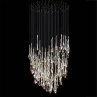 A striking custom lighting installation featuring a large chandelier with a cylindrical shape. This hotel chandelier comprises numerous crystal-like glass elements suspended at varying lengths on thin wires, creating a cascading effect. The glass pieces range from elongated drops to twisted, abstract forms, each reflecting and refracting light brilliantly. The lower portion of the chandelier is densely packed with these illuminated glass elements, emitting a warm, sparkling glow. Above the main crystal cluster, the thin wires extend upward, terminating in small LED lights that form a dome-like canopy, resembling a starry night sky. This big chandelier offers a perfect blend of contemporary design and classic luxury, ideal for creating a dramatic focal point in upscale spaces. The contrast between the bright, twinkling lower section and the subtle, star-like upper portion adds depth and visual interest to the overall design.