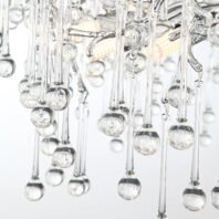 A close-up view of a modern glass chandelier featuring numerous hanging glass elements. The image shows multiple thin, elongated glass rods of varying lengths, each terminating in a small spherical glass droplet. These droplets appear to contain tiny air bubbles or have a slightly frosted texture, giving them a subtle, ethereal quality. The glass rods and droplets are suspended from what seems to be a more complex structure of intertwining glass branches, partially visible at the top of the image. In the background, a warm, golden light source is visible, likely an LED or other modern lighting element with a honeycomb or textured diffuser. This light illuminates the glass elements from within, creating a sparkling effect and highlighting the intricate design of the chandelier. The overall effect is one of elegance and modernity, with the chandelier resembling a crystalline rain shower or an abstract interpretation of icicles.