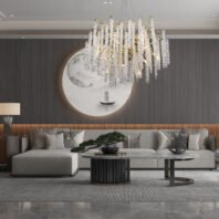 A luxurious living room featuring a large crystal and gold chandelier as the centerpiece. The room has a modern Asian-inspired design with a gray wood-paneled wall, adorned with a circular backlit artwork depicting a minimalist landscape with a boat. A plush L-shaped sofa in light gray occupies the foreground, accented with various throw pillows and a small bonsai tree. Two modern coffee tables with dark bases and light tops sit in front of the sofa. The floor is covered with a textured gray rug, and recessed lighting in the ceiling complements the chandelier's warm glow. The overall aesthetic blends contemporary luxury with subtle Asian influences.