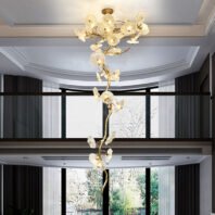 A striking, vertically elongated chandelier featuring glass flower-like elements on a golden branch, spanning two floors of an elegant interior. The chandelier resembles a tall, artistic tree with illuminated glass blooms. Its golden stem twists upwards, adorned with numerous translucent, textured glass shades that resemble flowers or sea anemones. Each glass element houses a light, creating a warm, ethereal glow. The fixture is suspended from a white coffered ceiling on the upper floor and extends down through an opening to the lower floor. The room has a modern design with dark curtains framing large windows that offer a view of trees outside. The chandelier serves as a stunning centerpiece, connecting the two levels visually and adding a touch of organic, nature-inspired luxury to the space.