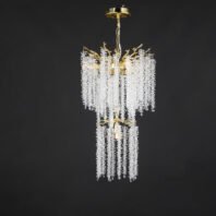 Luxurious two-tier chandelier with cascading crystal strands hanging from gold-toned metal frame. Features organic branch-like design, gold canopy, and chain. Crystal droplets create a waterfall effect against dark background. Warm lighting illuminates the piece.