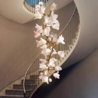 Exquisite branch chandelier hangs gracefully in a stairwell, featuring delicate white and pink flower-shaped lights. Golden branches intertwine, supporting numerous illuminated blooms that cascade downward. The chandelier complements a curved marble staircase with sleek metal railings and gold accents. This stunning branch chandelier creates a focal point in the neutral-toned interior, blending nature-inspired design with modern luxury. Soft, warm light from the floral fixtures adds a romantic ambiance to the elegant space, showcasing the chandelier's artistry against the sweeping lines of the staircase.