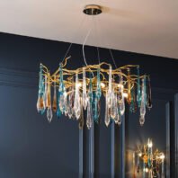 Elegant chandelier with golden branches and hanging glass icicles in clear, blue, and amber colors. Illuminated bulbs cast a warm glow. Suspended from a circular mount against a dark blue wall with decorative molding. A smaller, matching chandelier is partially visible in the background.