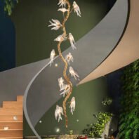 A modern interior featuring a striking vertical chandelier shaped like a golden branch with illuminated glass birds. The chandelier hangs alongside a sleek, curved white staircase with wooden steps. The background is a deep green, creating a natural ambiance. Lush plants are visible in the lower right corner, enhancing the nature-inspired theme. The combination of the organic chandelier design, the smooth lines of the staircase, and the greenery creates a harmonious blend of contemporary architecture and natural elements.