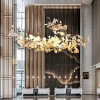 A grand hotel lobby featuring a stunning, elongated chandelier with illuminated leaf-like shapes. The fixture hangs above reception desks, casting a warm glow. Behind, a dramatic wall combines metallic curtains and dark panels with hanging pendant lights. Geometric patterns on glass partitions frame the space. The design blends organic forms with modern luxury, creating an impressive and welcoming entrance area.