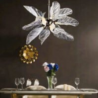 A stunning glass leaf chandelier hangs above a set dining table. The chandelier features translucent leaves with intricate vein patterns, resembling a blooming flower. Below, a table is elegantly set with wine glasses, plates, and a vase of blue and pink roses. A decorative gold sunburst wall ornament is visible in the background. The scene combines natural elements with modern luxury, creating a sophisticated dining atmosphere.