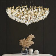 An opulent gold and crystal chandelier hangs above a white round table. The chandelier features intricate gold branches adorned with numerous teardrop-shaped crystal pendants. Below, a vase with pampas grass and white ceramic decorative objects sit on the table. The backdrop is a dark wall, creating striking contrast with the illuminated chandelier.