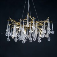 An elegant and contemporary chandelier suspended against a dark background. The fixture features a central structure of intertwining gold-plated branches that form an organic, tree-like silhouette. From these golden branches hang numerous clear glass pendants, each with a unique, elongated shape reminiscent of icicles or abstract flowers. The glass elements have long, slender stems that transition into more elaborate, bulbous forms at their ends, creating a cascading effect. The chandelier is illuminated from within, with several visible LED lights nestled among the branches, casting a warm glow that highlights both the metallic gold tones and the translucent glass pendants. The light refracts and reflects through the glass elements, creating a dazzling display of sparkles and subtle rainbow effects. The piece is suspended by thin wires from the ceiling, giving it a floating appearance. The contrast between the warm gold of the branches and the cool, clear glass of the pendants creates a striking visual balance. This chandelier beautifully combines natural, organic forms with luxurious materials and modern lighting technology, resulting in a piece that is both a functional light source and a stunning sculptural element. It would serve as an impressive focal point in contemporary or transitional interior spaces, particularly suited for dining areas, grand entryways, or upscale living rooms.