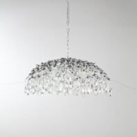 A stunning dome-shaped chandelier suspended against a light background. The fixture features a dense canopy of crystal elements arranged in a cascading, umbrella-like formation. The top of the dome is adorned with silver-toned metal elements shaped like leaves or vines, creating an organic, nature-inspired look. These metallic accents gradually give way to an abundance of clear crystal pendants hanging below. The crystal elements vary in size and shape, with larger teardrop-shaped crystals forming the outer rim of the chandelier. These crystals catch and refract light brilliantly, creating a dazzling, prismatic effect with hints of rainbow colors visible throughout the piece. The chandelier is suspended by a silver chain, complementing the silver leaf accents. Thin wires can be seen supporting the structure, adding to its delicate appearance. This luxurious lighting fixture combines the elegance of crystal with the organic beauty of silver foliage, resembling a frozen waterfall or a crystal-encrusted tree. It would serve as a captivating focal point in any upscale interior, suitable for dining rooms, entryways, or living spaces where it can showcase its full splendor.