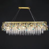 An opulent rectangular chandelier suspended against a dark background, showcasing a stunning combination of gold, clear crystals, and colorful gemstone-like accents. The fixture is framed by a polished gold rectangular structure, hanging from a matching gold ceiling mount via thin cables. From this frame, intricate gold branches extend downward, adorned with multi-colored crystal flowers in hues of blue, turquoise, amber, and clear. These vibrant crystal accents provide pops of color against the predominantly gold and clear crystal design. The most striking feature is the abundant cascade of clear crystal strands hanging from the gold frame, creating a shimmering curtain effect. These crystal strands are arranged in uniform lengths, giving the chandelier a waterfall-like appearance. The clear crystals catch and refract light, producing a dazzling sparkle. The chandelier is illuminated from within, causing the entire piece to glow warmly against the dark background. This lighting fixture masterfully combines the warmth of gold with the cool brilliance of crystals, resulting in a piece that is both a functional light source and a luxurious work of art, capable of making a dramatic statement in any high-end interior space, particularly suited for long dining tables or expansive areas.