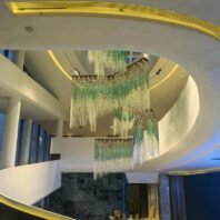 A stunning large chandelier showcasing custom lighting design in a luxury hotel lobby. This big chandelier features multiple tiers of cascading crystal-like elements in emerald green and clear, creating a mesmerizing waterfall effect. The hotel chandelier hangs gracefully from a curved ceiling with golden accents. Crystal strands shimmer and transition from vibrant green to clear, mimicking a lush forest canopy. This exquisite custom lighting installation transforms the modern, multi-level lobby into an enchanting, nature-inspired environment.