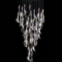 A striking large chandelier showcasing innovative custom lighting design for upscale hotels. This big chandelier features numerous translucent glass elements shaped like elongated leaves or petals, suspended at varying heights on thin wires to create a dramatic cascading effect. Each leaf-like piece is illuminated from within, emitting a soft, ethereal glow. The hotel chandelier's unique composition resembles a waterfall of light or a cluster of floating leaves. Small LED lights at the top of each wire add extra sparkle and depth, creating a mesmerizing play of light and shadow. This exquisite custom lighting installation transforms the space into an enchanting, nature-inspired display, perfect for creating a sophisticated and luxurious atmosphere in high-end hospitality environments.