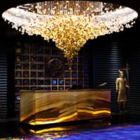 A spectacular large chandelier showcasing innovative custom lighting design for upscale hotels. This big chandelier features a circular base with thousands of golden and crystal elements cascading downward, creating a stunning inverted dome effect. The hotel chandelier's center glows with warm golden light, transitioning to clear crystals at the edges. Below, an illuminated onyx reception desk adds to the luxurious ambiance. The backdrop includes a decorative wall with intricate patterns and a golden statue, exemplifying how this exquisite custom lighting transforms high-end hospitality spaces into opulent, eye-catching environments.