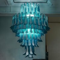 A stunning large chandelier showcases innovative custom lighting design for upscale hotels. This big chandelier features multiple tiers of alternating teal blue and white glass elements, creating a mesmerizing waterfall-like effect. The hotel chandelier's unique composition resembles cascading ocean waves or layered ice formations. The glass pieces are elongated and curved, arranged in a circular pattern that narrows towards the bottom. Illuminated from within, the chandelier emits a soft, ethereal aqua glow that highlights the interplay between the transparent teal and opaque white elements. This bespoke lighting installation transforms the space into an artistic underwater-inspired display, perfect for elevating high-end hospitality environments with its blend of organic design and sophisticated illumination.