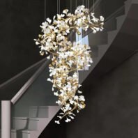 A stunning spiral chandelier featuring white glass petals and gold-toned branches, suspended in a modern stairwell. The chandelier has an organic, tree-like structure that twists downward in a graceful helix. Numerous white glass elements, resembling stylized flowers or leaves, are attached to the golden metallic framework, creating a cascading effect. The fixture is illuminated, casting a warm glow that contrasts beautifully with the dark walls of the stairwell. A portion of a modern staircase with light-colored steps and handrails is visible in the background, its angular lines providing a striking contrast to the chandelier's fluid form. The chandelier's intricate design and the interplay between the white glass and gold metal create a luxurious and eye-catching focal point in this contemporary interior space, bridging the gap between artistic sculpture and functional lighting.