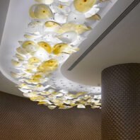 A breathtaking large chandelier showcases innovative custom lighting design for luxury hotels. This big chandelier features a stream of suspended glass or acrylic petal-like shapes in white and soft yellow hues, flowing along a curved ceiling. The hotel chandelier creates a dynamic, organic form that contrasts beautifully with the geometric patterns of the textured walls. Delicate wire suspensions give the illusion of floating petals. This unique lighting installation transforms the corridor into an artistic display of light and movement, demonstrating how bespoke illumination can elevate high-end hospitality environments.