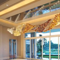 An exquisite custom lighting hotel chandelier adorns a spacious lobby with floor-to-ceiling windows overlooking a lush tropical landscape. The chandelier features a flowing, wave-like design composed of thousands of glass elements in various golden hues, transitioning from clear to light gold, deep amber, and rich bronze. The undulating form creates a sense of fluid movement across the ceiling. Above the main chandelier, a recessed area with additional lighting elements adds depth and a sky-like effect. The warm, golden glow of the chandelier contrasts beautifully with the cool, natural scenery outside, which includes palm trees, a manicured lawn, and a glimpse of a pool. The polished floor reflects the chandelier's brilliance, enhancing the overall luxurious atmosphere of the space and creating a seamless blend between interior opulence and exterior paradise.